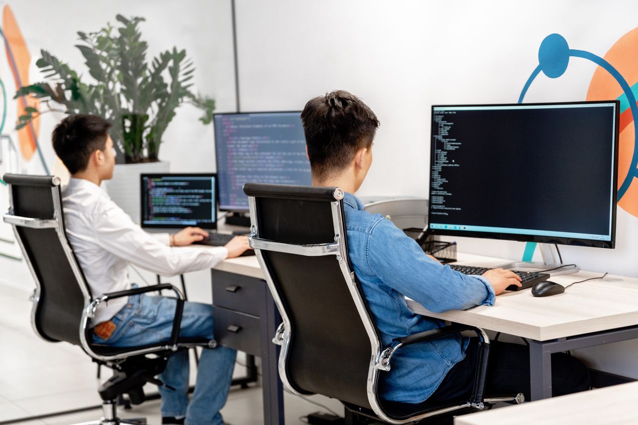 Home Two male developers at desks programming in a modern office workspace with large monitors.